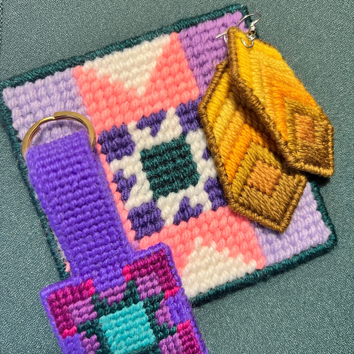 A set of handmade crocheted keychains and a matching square patch laid on a teal-green textured surface. The set includes a purple keychain with a gold ring and a matching geometric patch, a yellow and gold geometric keychain, and a larger square patch with a pink, purple, white, and teal geometric pattern.