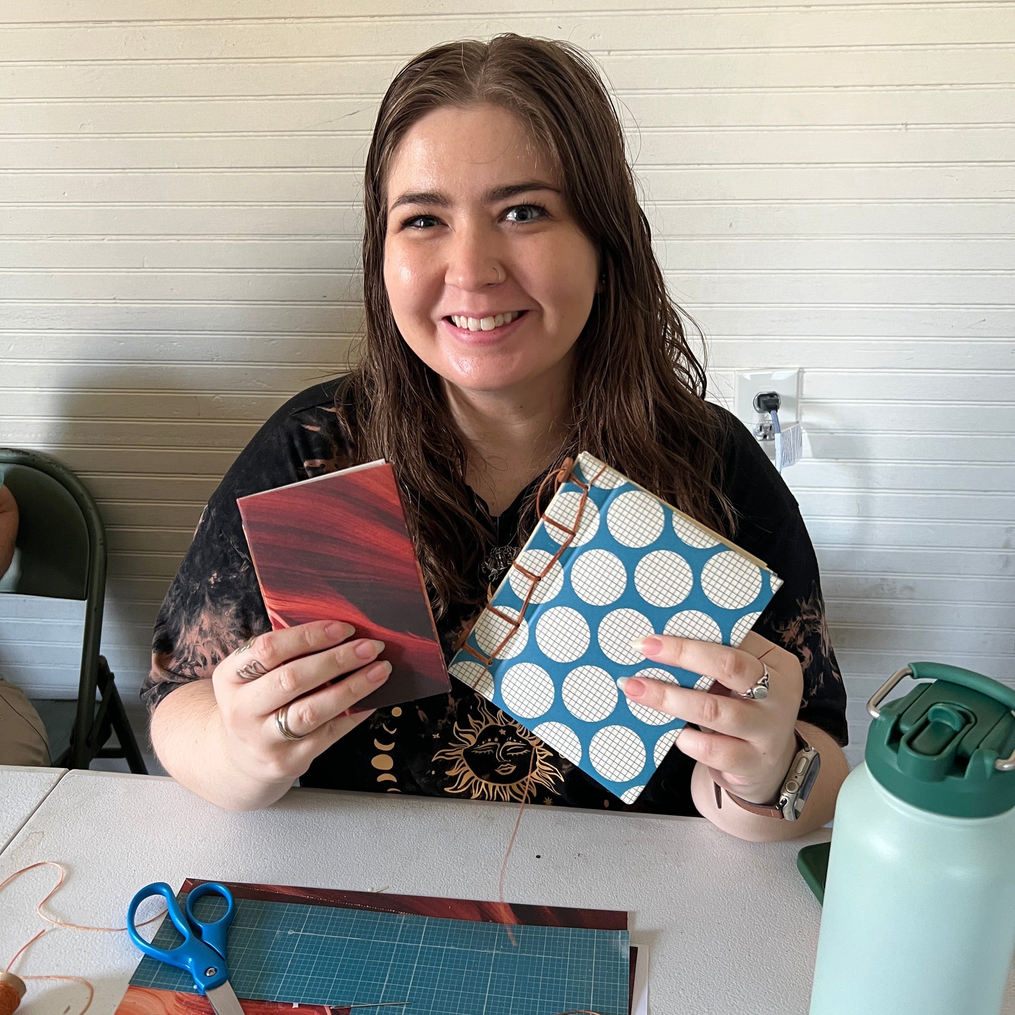 A smiling woman with long brown hair sits at a table holding two small handmade books or notebooks. One has a red and black marbled cover, and the other has a blue background with white polka dots.