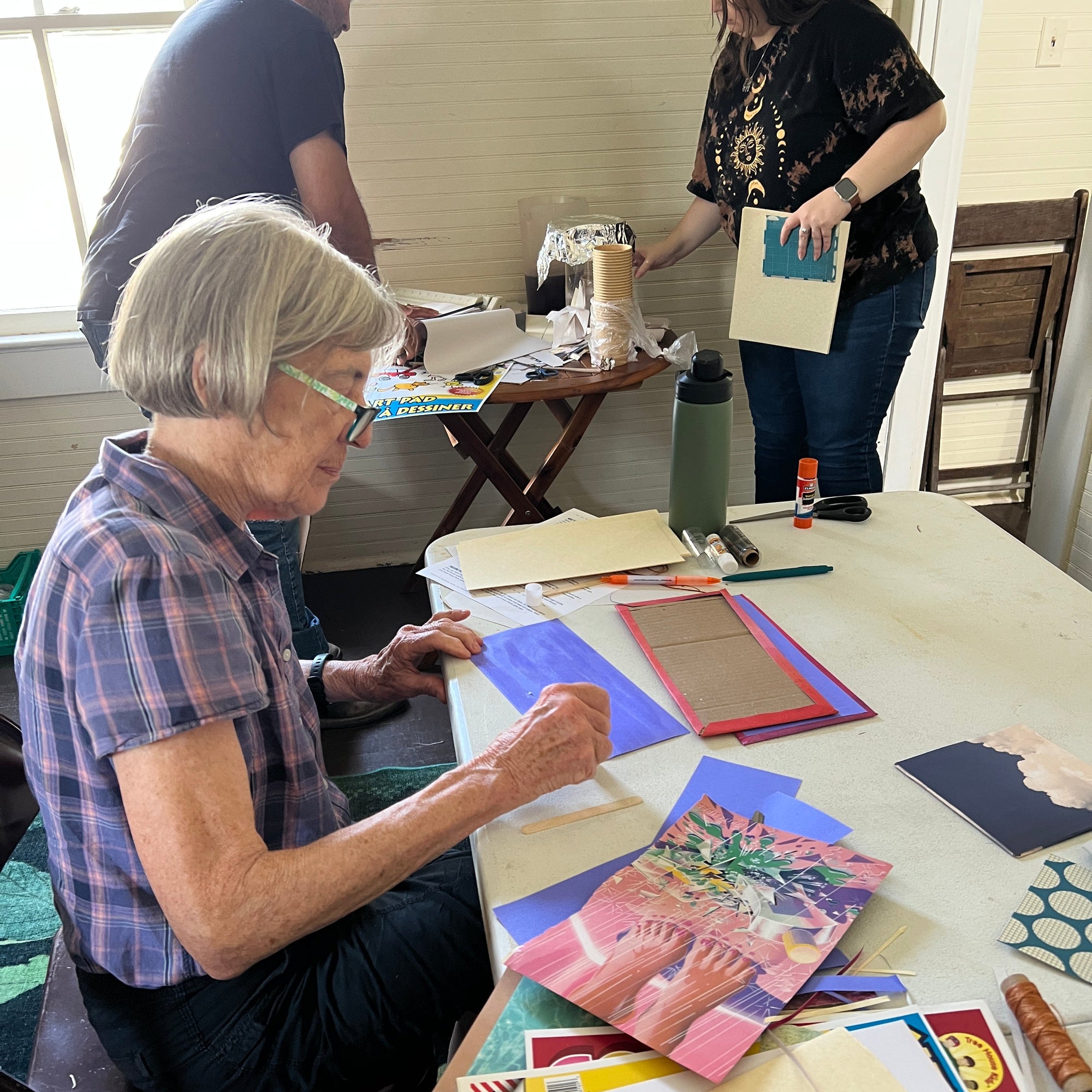 A woman with short gray hair and glasses sits at a table, working on a colorful art project with paper, fabric, and printed materials. Two other people are visible in the background and more crafting supplies in a home studio setting.