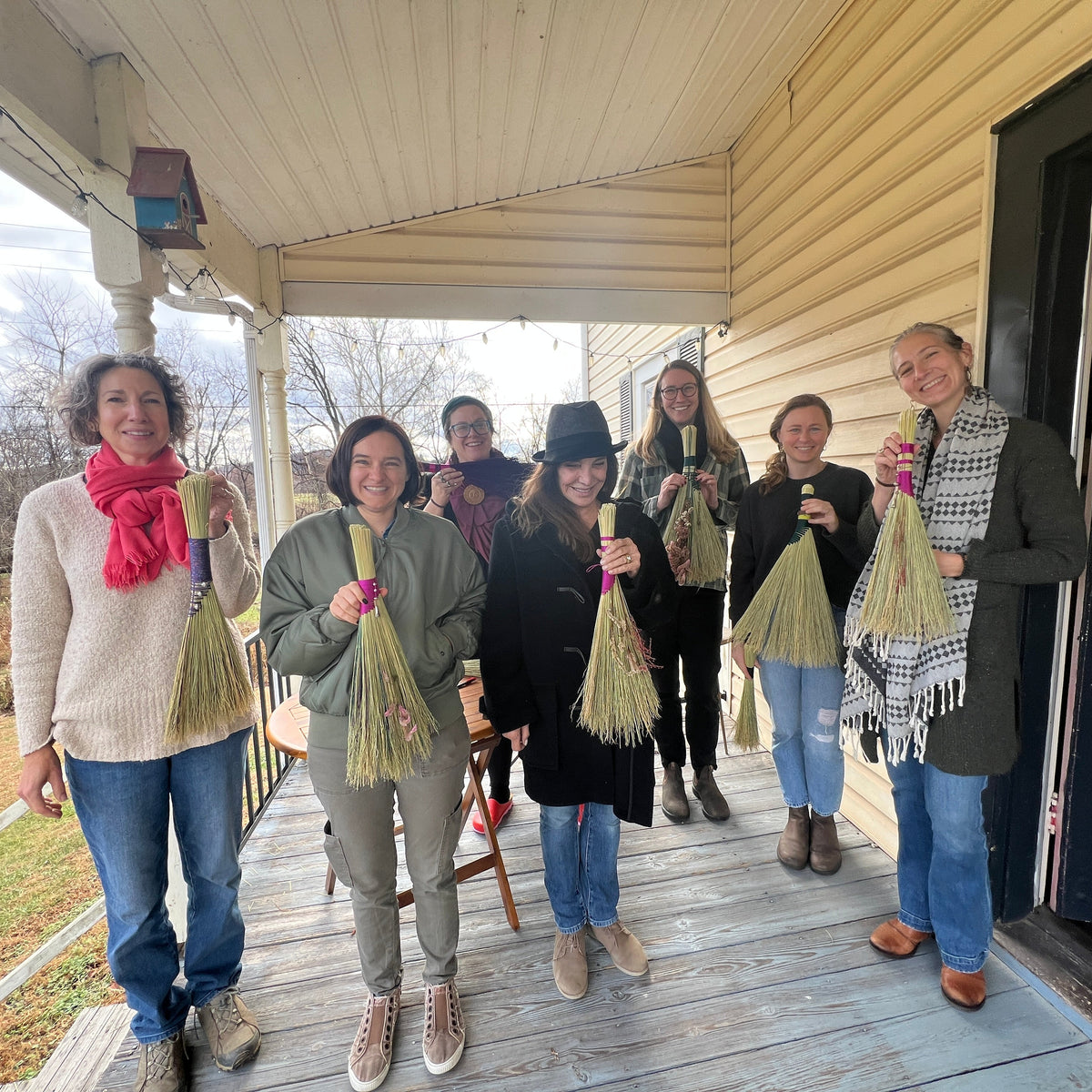 A group of six women standing on a wooden porch holding handmade straw brooms. The women are dressed in casual autumn clothing, including scarves and jackets, and are smiling and posing together on the porch of a house with beige siding.