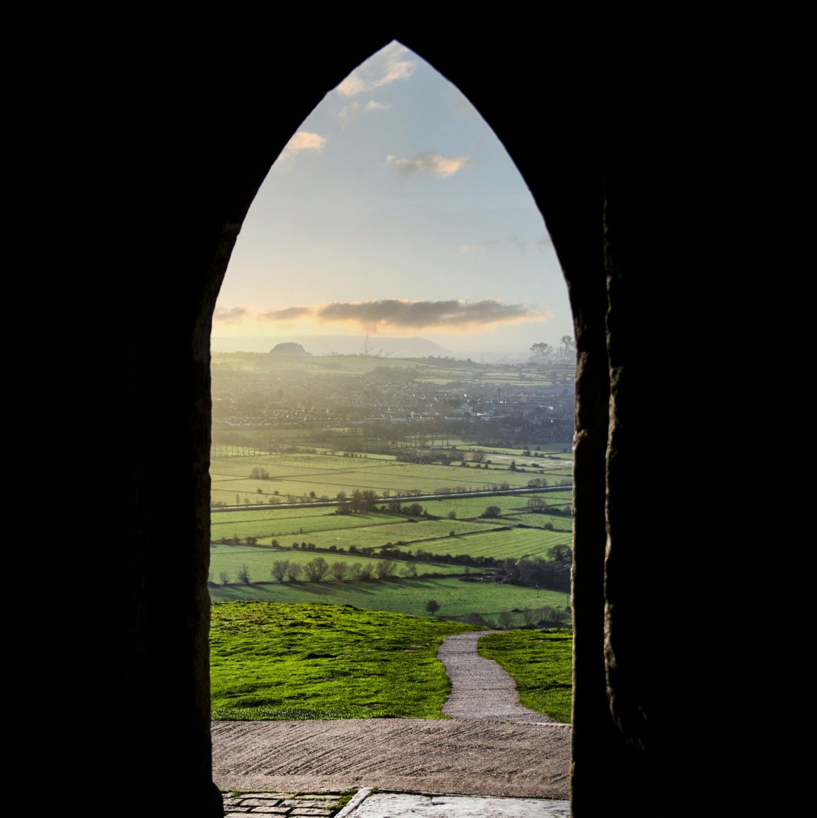 View through a stone archway of a landscape with green fields and a path.